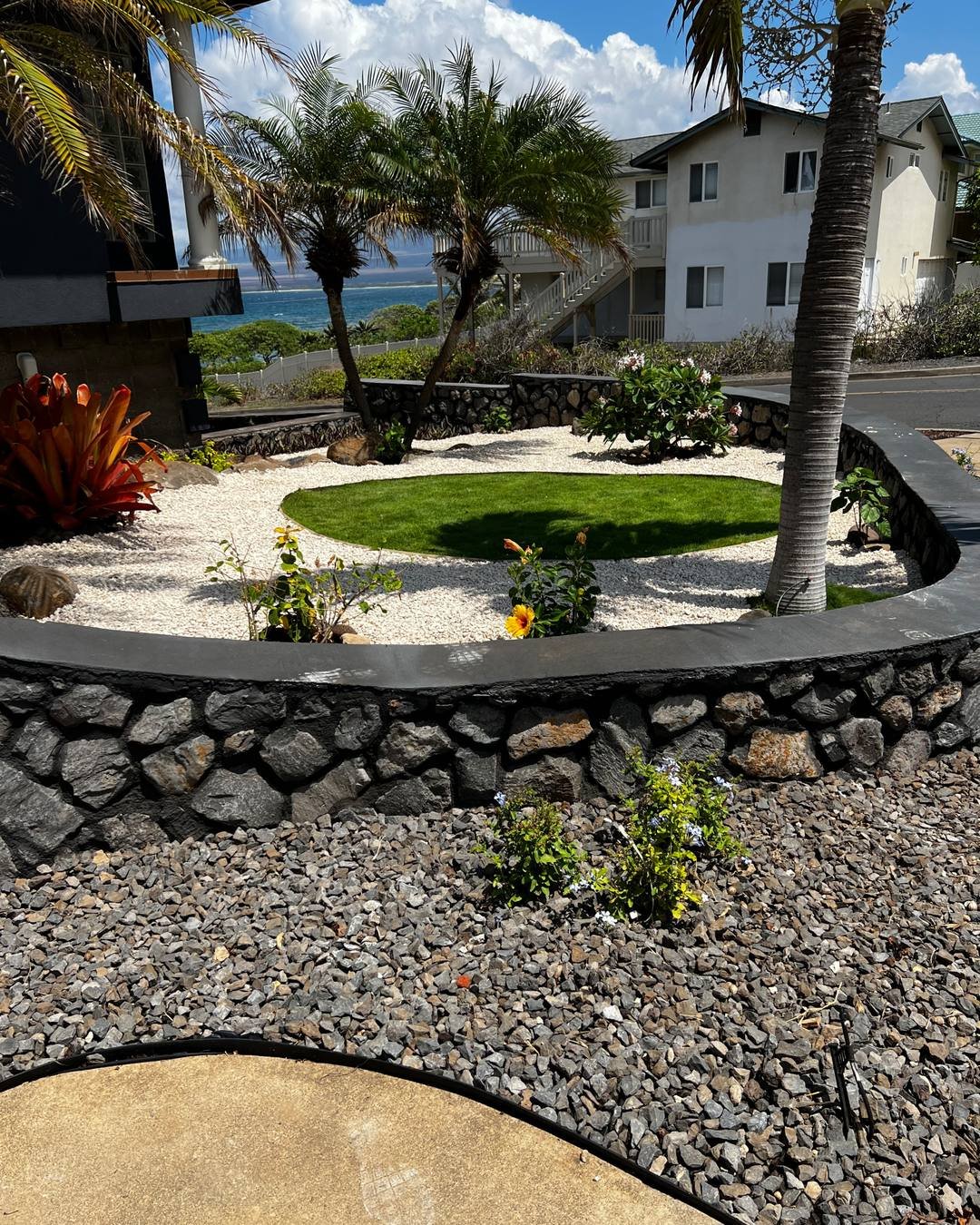 A finished Maui front yard by Roby Landscape: a curved stacked lava rock wall with a smooth concrete cap holding back a bed of white gravel, an oval of green lawn, palms and a red bromeliad, with the ocean behind the house.