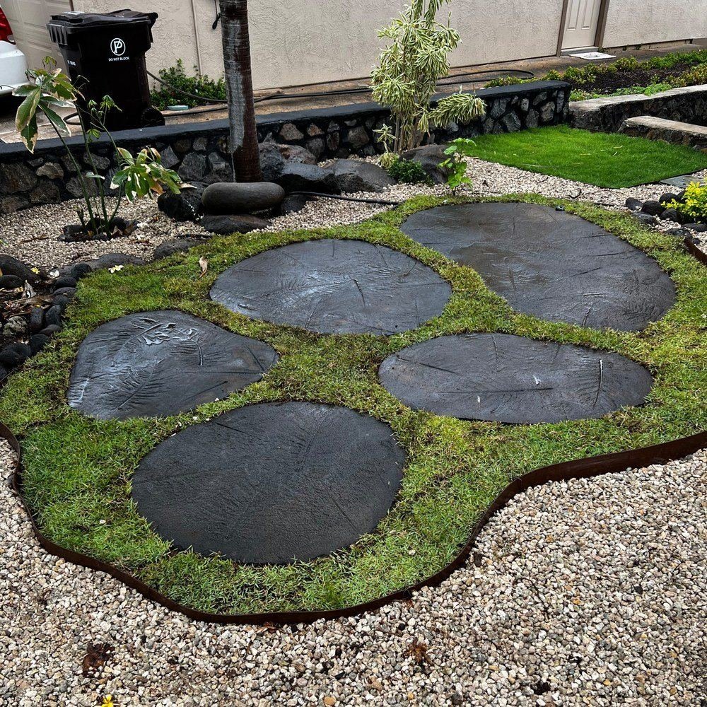 Round dark stepping stones set flush into a green turf island edged in steel, surrounded by pale gravel, with a stacked lava rock wall behind.